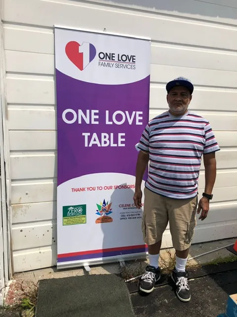 A man standing beside the One Love Table banner