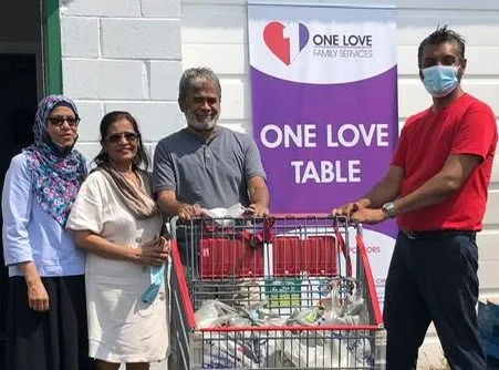 Four people with a shopping cart full of groceries beside the One Love Table banner