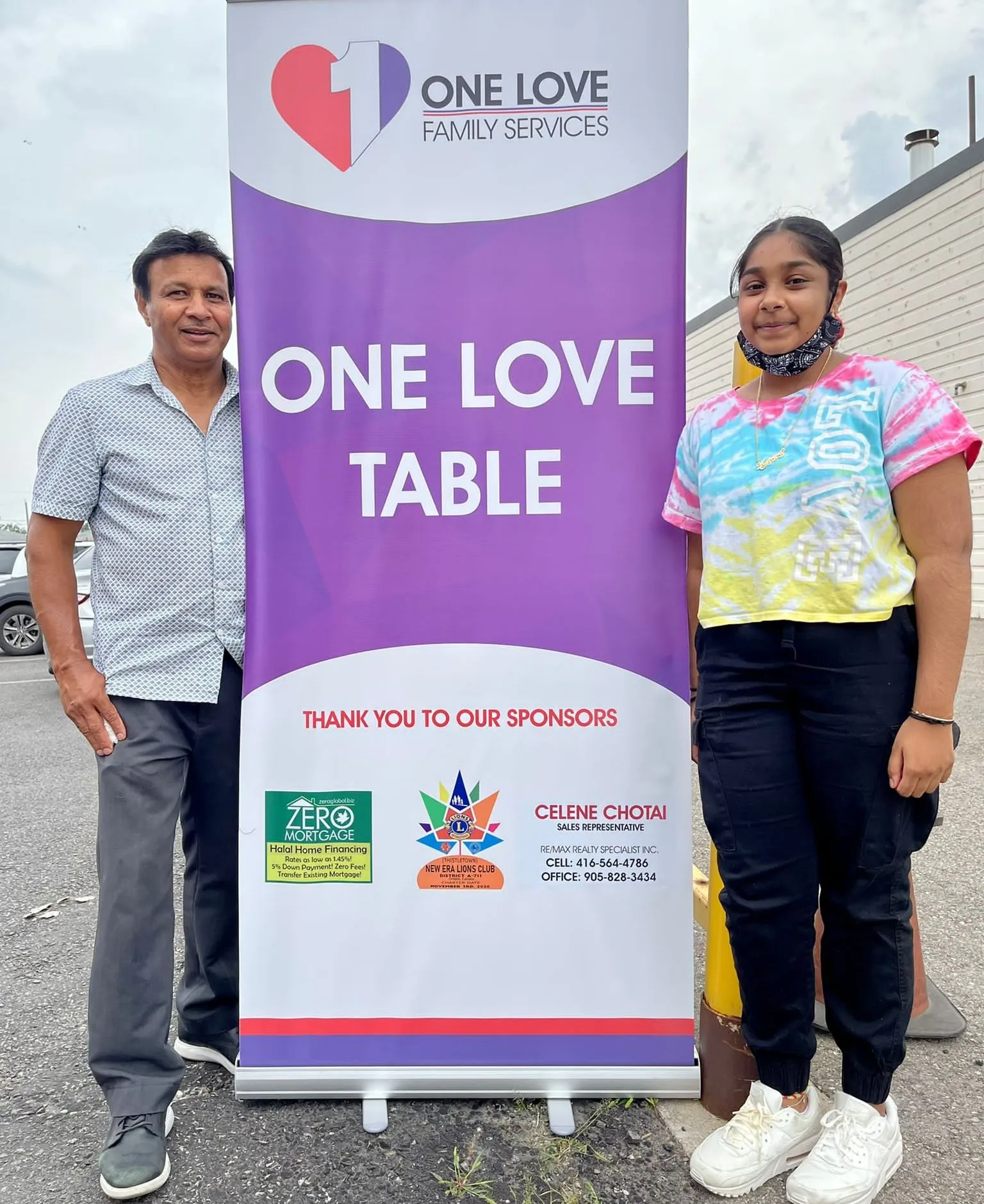 Two people smiling beside the One Love Table banner at a hamper distribution day